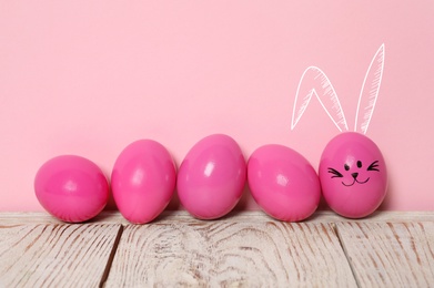 One egg with drawn face and ears as Easter bunny among others on white wooden table against pink background