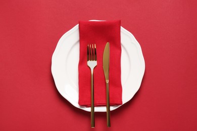 Photo of Clean plate with golden cutlery on red background, top view