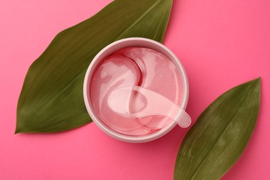 Photo of Jar of under eye patches with spoon and green leaves on pink background, flat lay. Cosmetic product