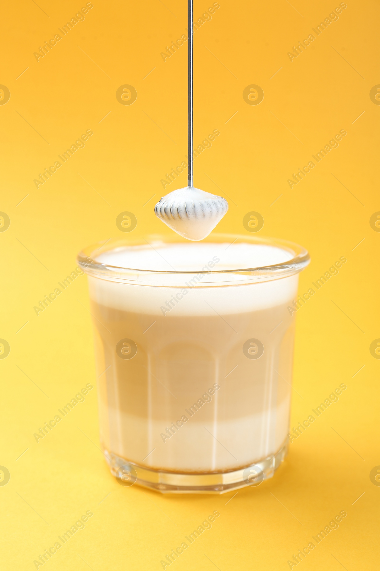 Photo of Mini mixer (milk frother) above tasty cappuccino in glass on yellow background