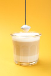 Photo of Mini mixer (milk frother) above tasty cappuccino in glass on yellow background