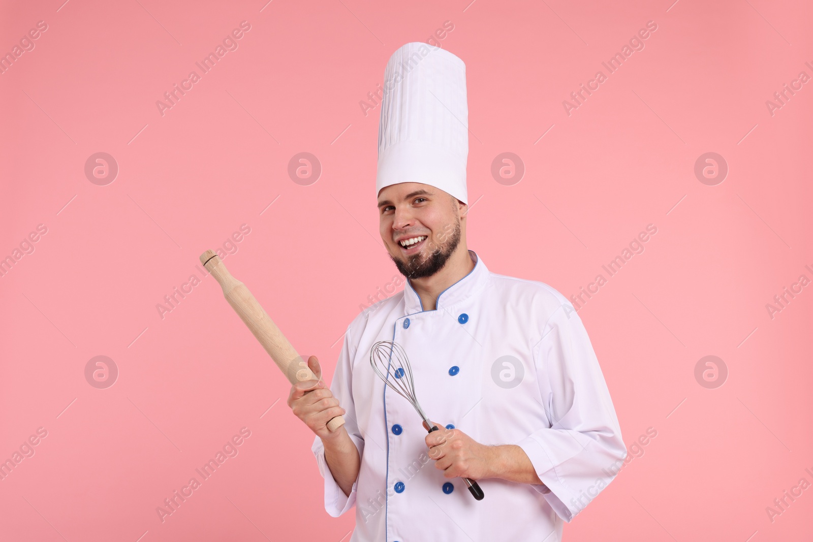 Photo of Happy professional confectioner in uniform holding rolling pin and whisk on pink background