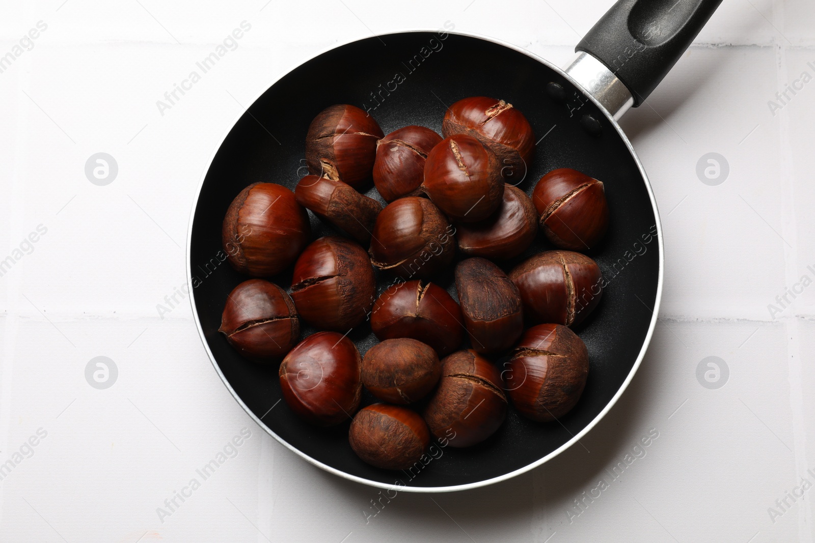 Photo of Fresh edible sweet chestnuts in frying pan on white tiled table, top view