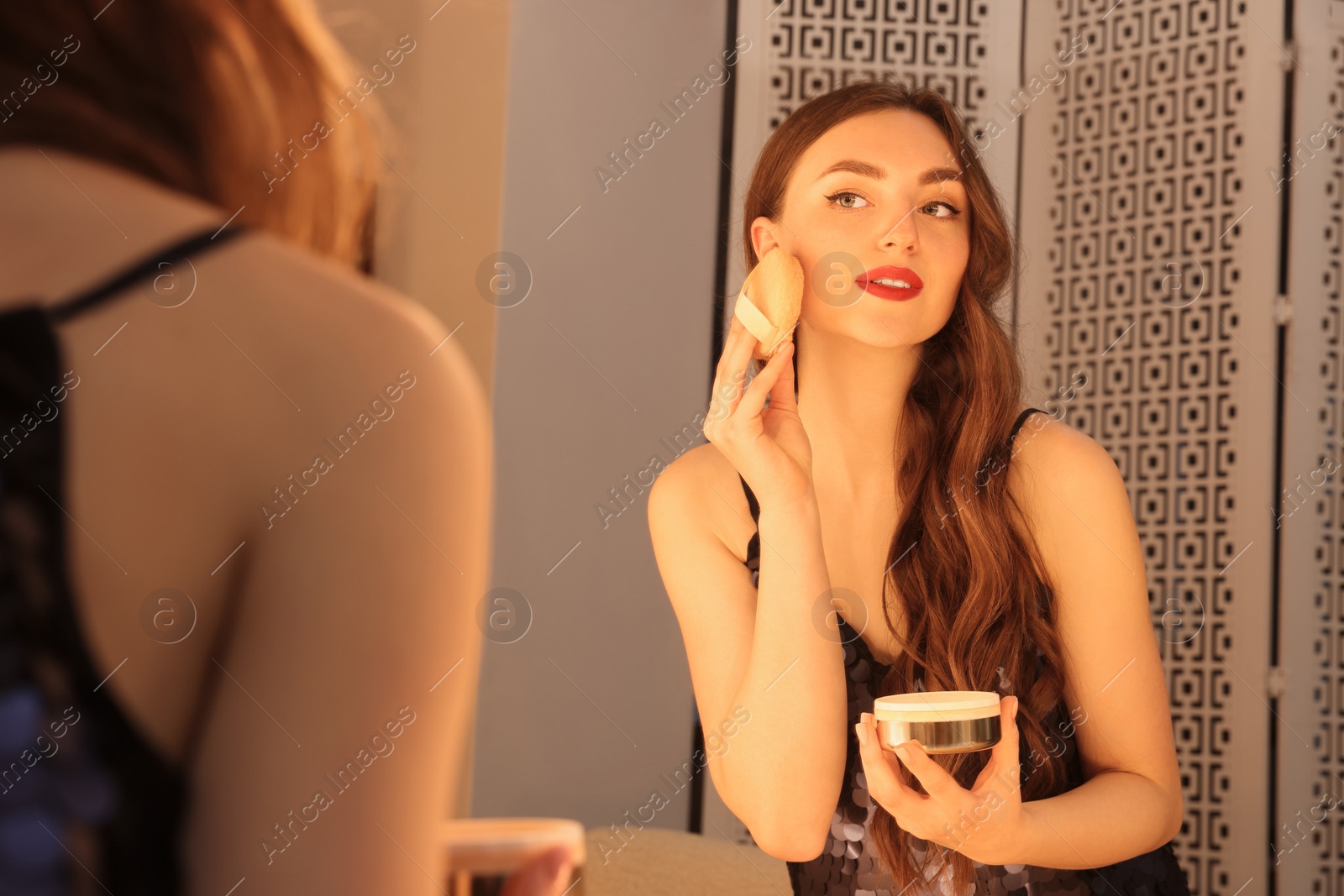 Photo of Beautiful young woman in elegant dress applying powder with puff applicator near mirror indoors