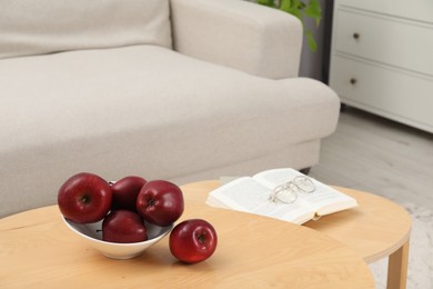 Red apples, book and glasses on wooden nesting tables near beige sofa indoors