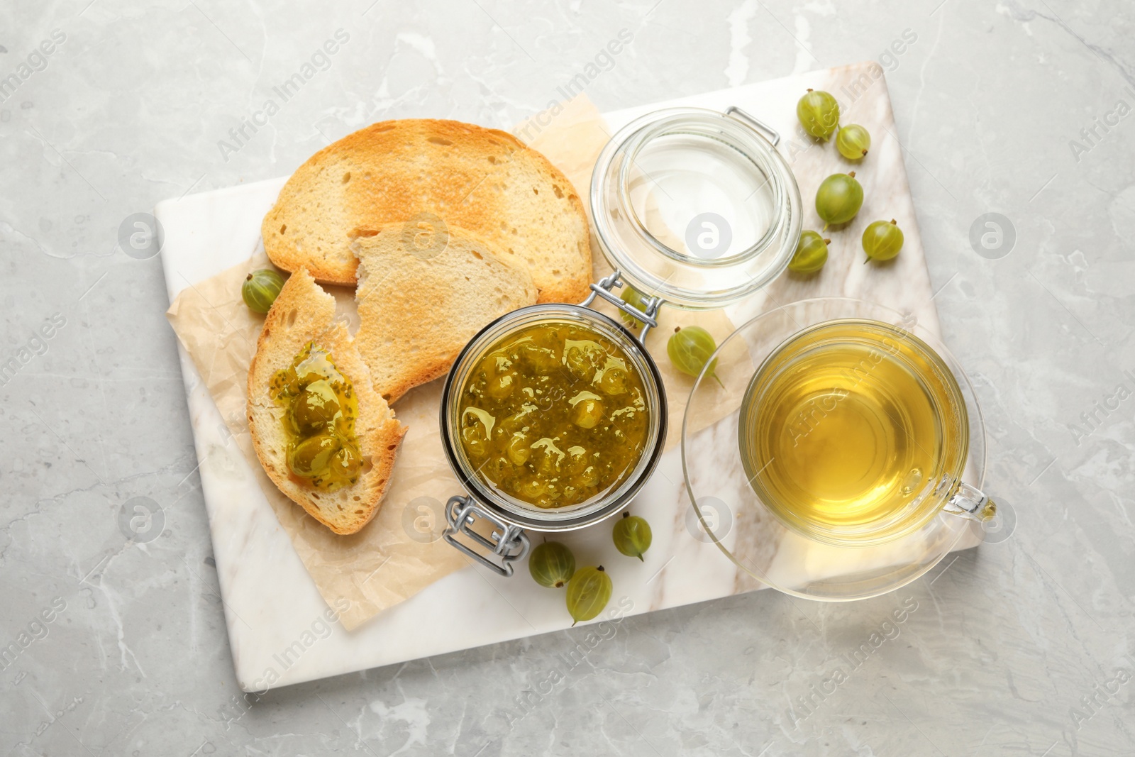 Photo of Jar of delicious gooseberry jam, bread, fresh berries and tea on grey marble table, top view