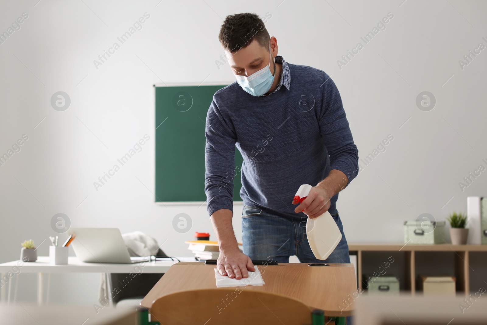 Photo of Teacher with protective mask disinfecting desk in classroom. Reopening after Covid-19 quarantine