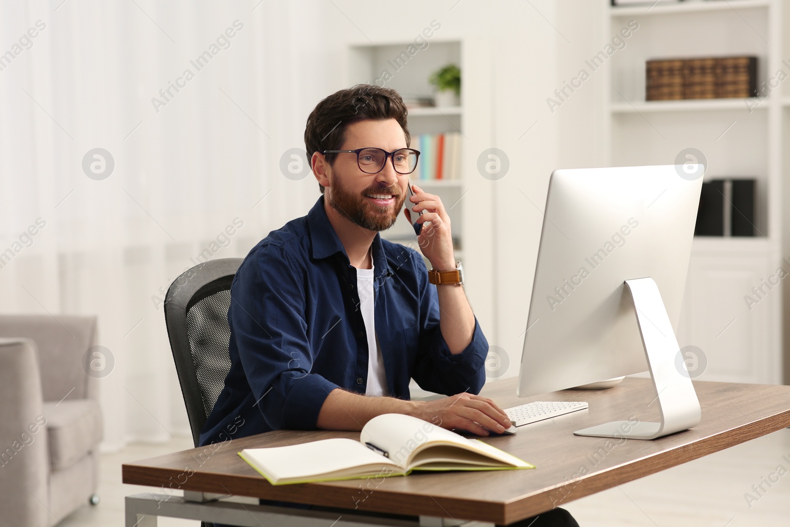 Photo of Home workplace. Happy man talking on smartphone while working with computer at wooden desk in room