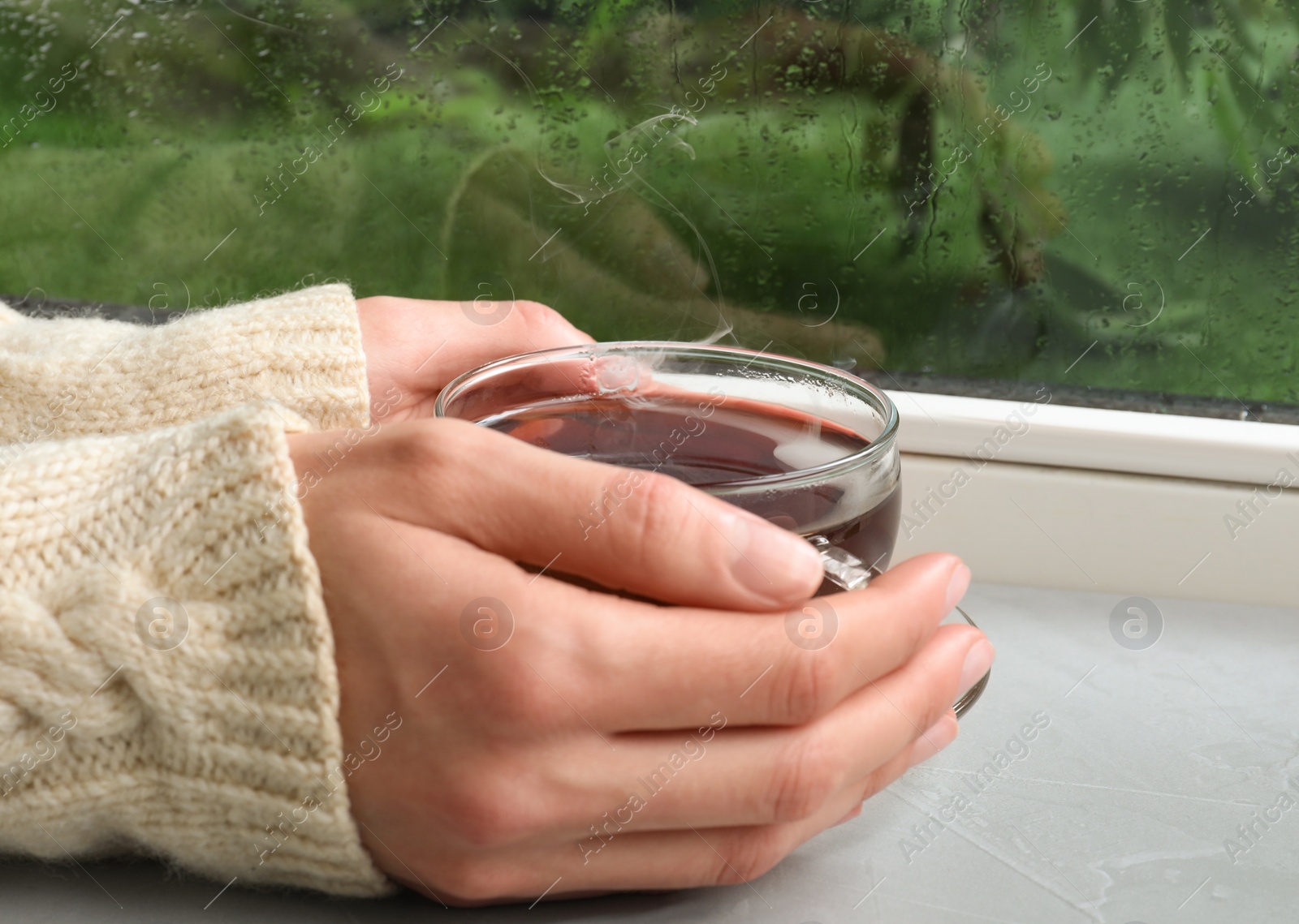 Photo of Woman with cup of hot tea near window on rainy day, closeup