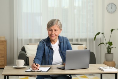 Photo of Beautiful senior woman taking notes while using laptop at wooden table indoors