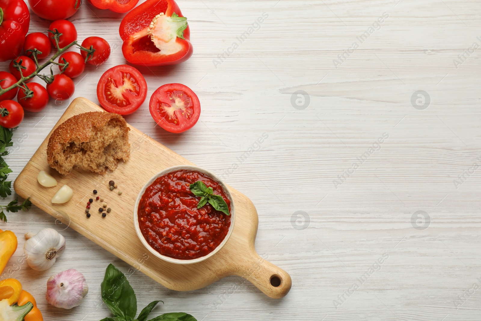 Photo of Delicious adjika sauce in bowl with bread and ingredients on white wooden table, flat lay. Space for text