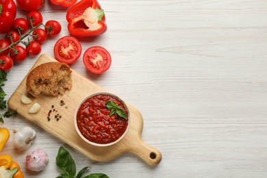 Delicious adjika sauce in bowl with bread and ingredients on white wooden table, flat lay. Space for text