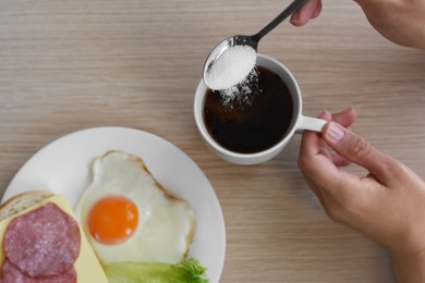 Woman adding sugar to morning coffee while having breakfast at wooden table, top view