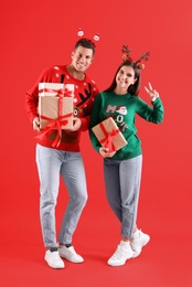 Photo of Beautiful happy couple in Christmas headbands and sweaters holding gifts on red background