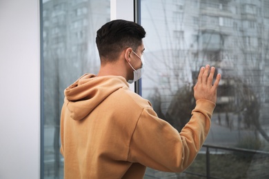 Photo of Sad man in protective mask near window indoors. Self-isolation during coronavirus pandemic