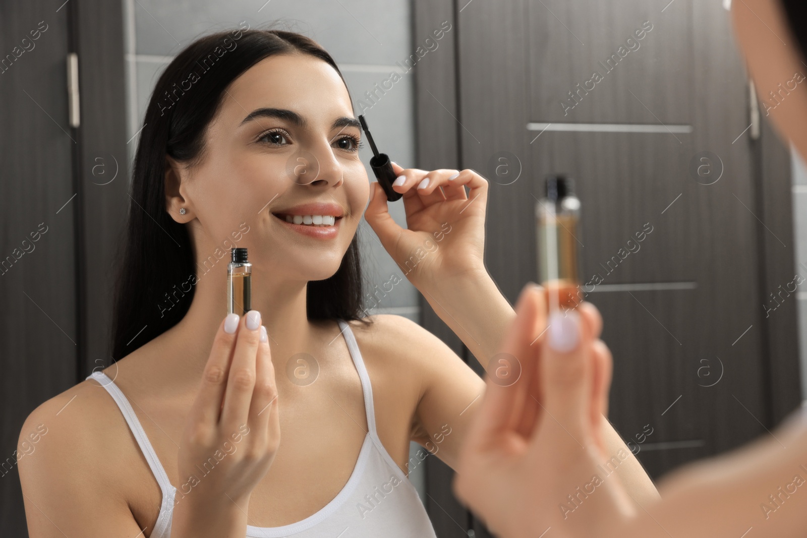 Photo of Young woman applying oil onto eyelashes near mirror indoors