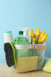 Photo of Basket with different cleaning supplies and beautiful spring flowers on wooden table against light blue background