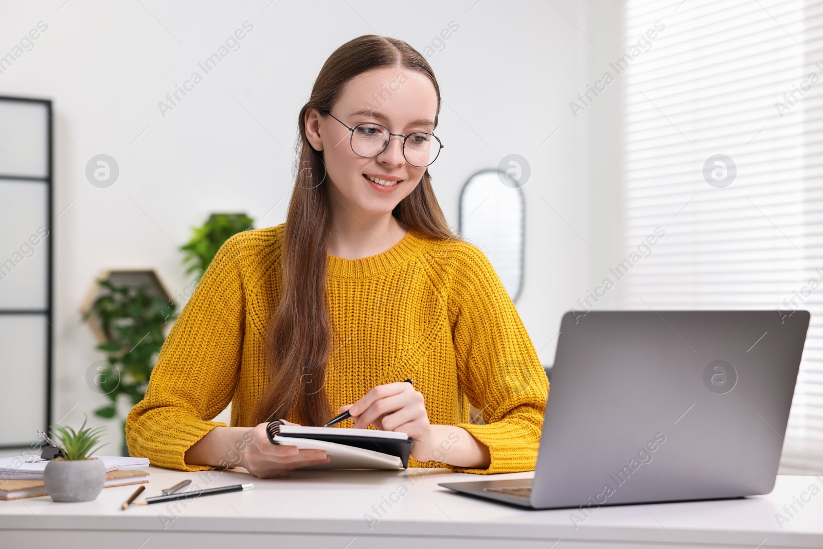 Photo of E-learning. Young woman taking notes during online lesson at white table indoors