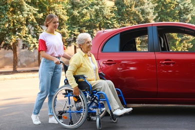 Photo of Senior woman in wheelchair with granddaughter near car outdoors