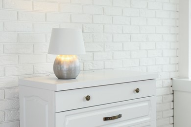 Photo of Modern chest of drawers with lamp near white brick wall