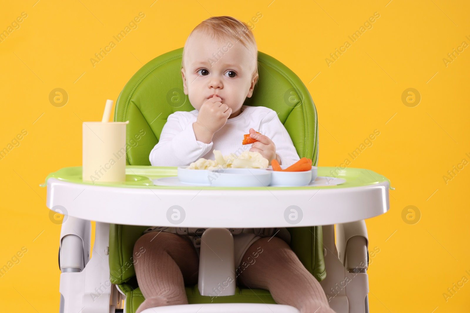 Photo of Cute little baby eating healthy food in high chair on orange background