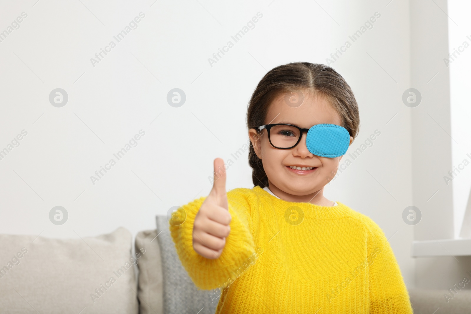 Photo of Happy girl with nozzle on glasses for treatment of strabismus showing thumbs up in room. Space for text