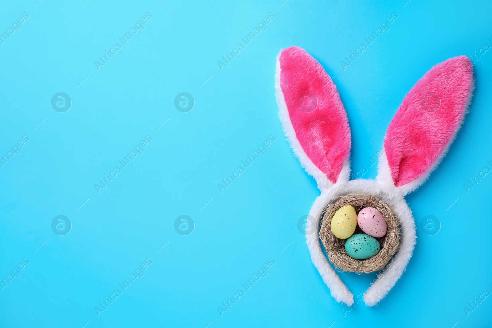 Photo of Headband with Easter bunny ears and dyed eggs in nest on light blue background, flat lay. Space for text