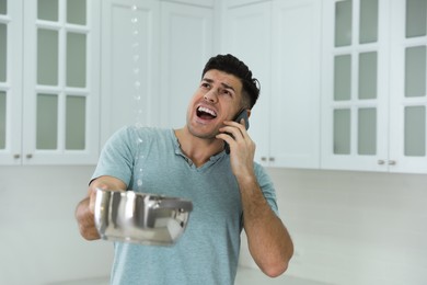 Photo of Emotional man calling roof repair service while collecting leaking water from ceiling in kitchen