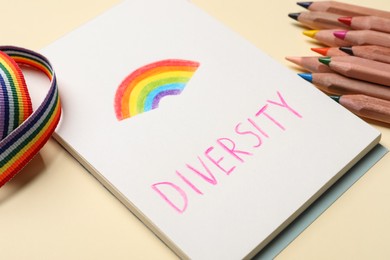 Photo of Paper sheet with word Diversity and drawn rainbow, pencils, ribbon on beige background