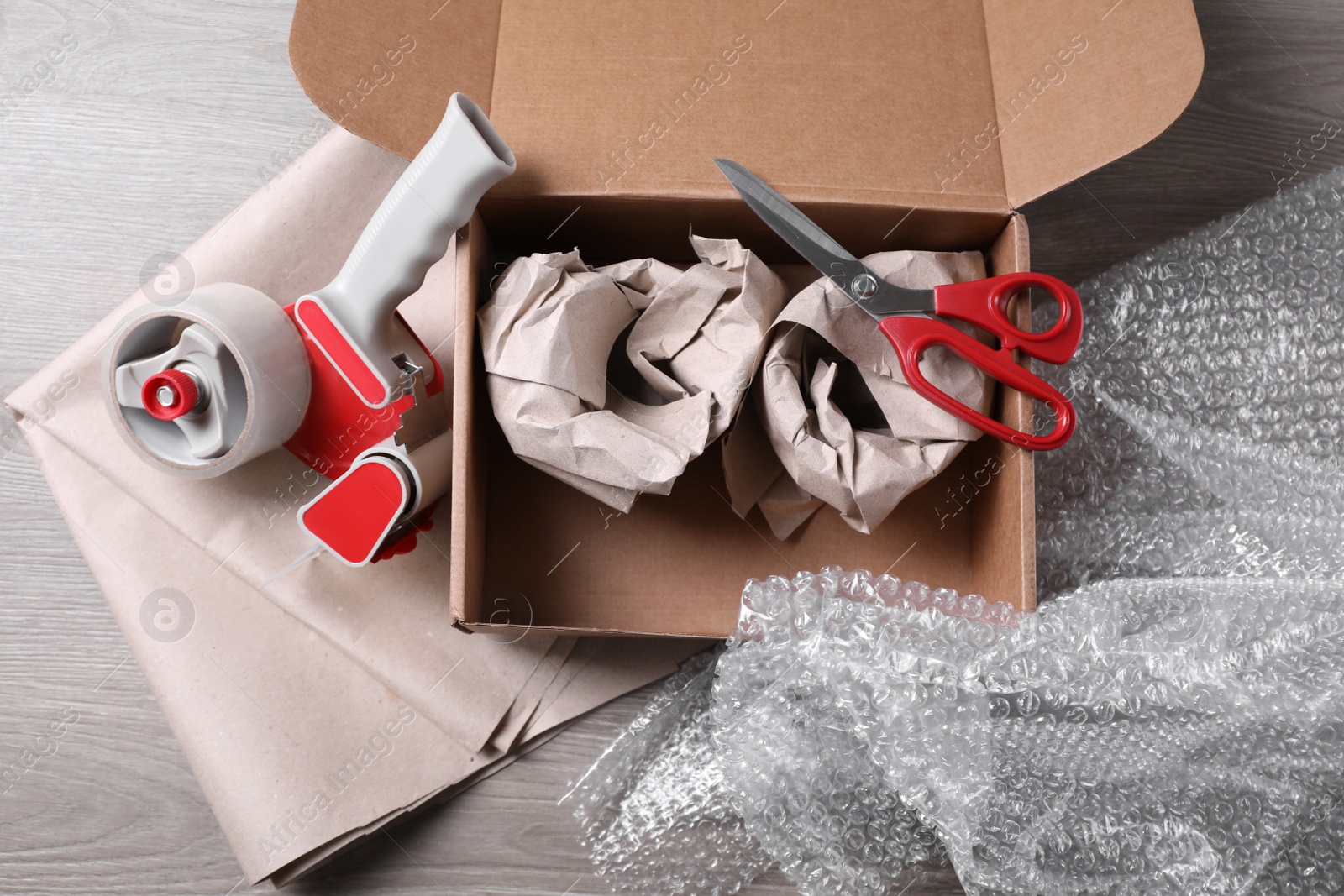 Photo of Open box with wrapped items, adhesive tape, scissors, paper and bubble wrap on wooden table, flat lay