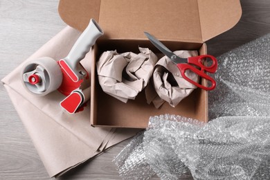 Photo of Open box with wrapped items, adhesive tape, scissors, paper and bubble wrap on wooden table, flat lay