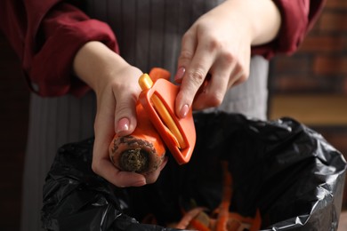 Photo of Woman peeling fresh carrot above garbage bin indoors, closeup