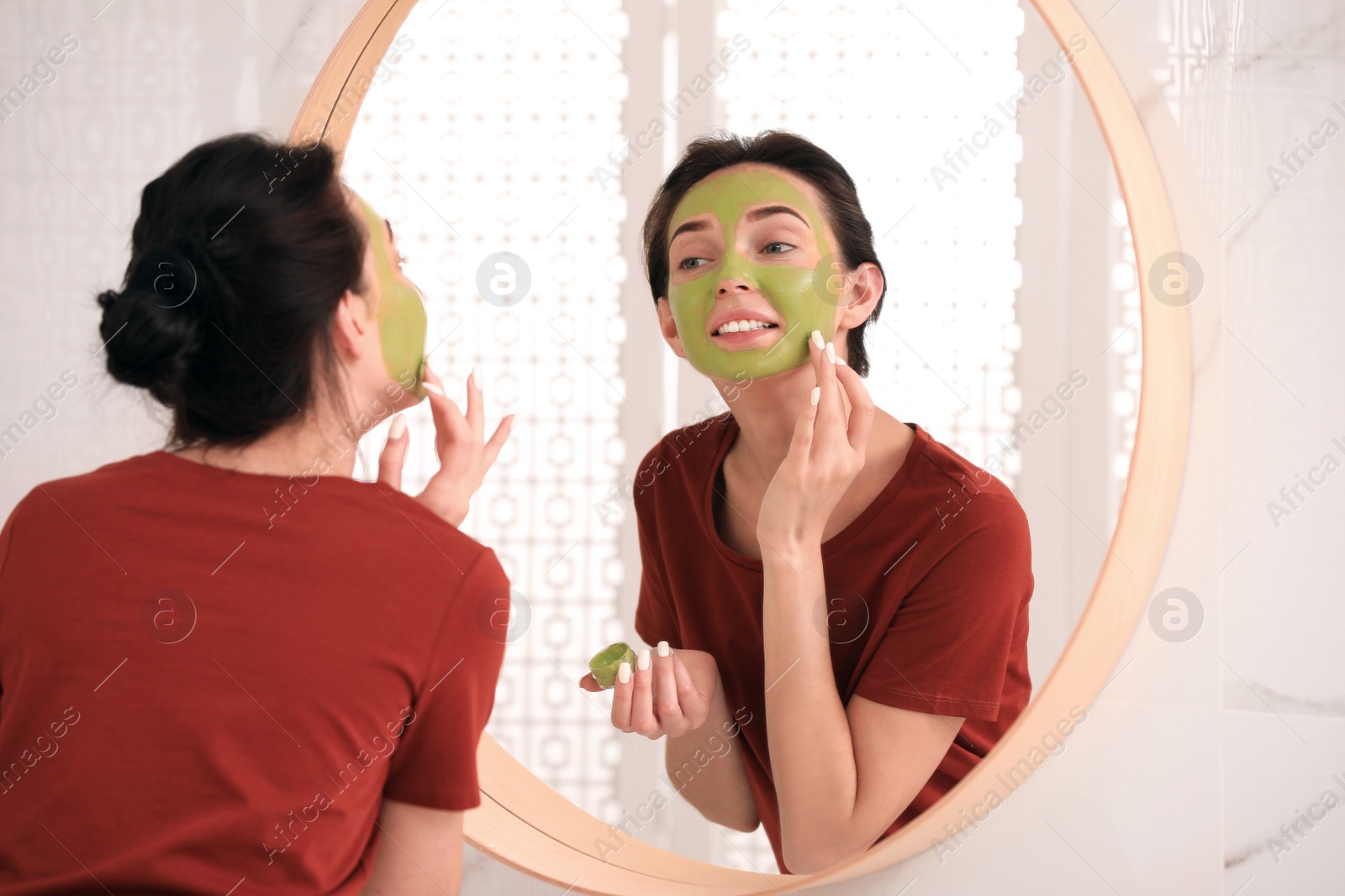 Photo of Young woman applying mask on her face near mirror in bathroom