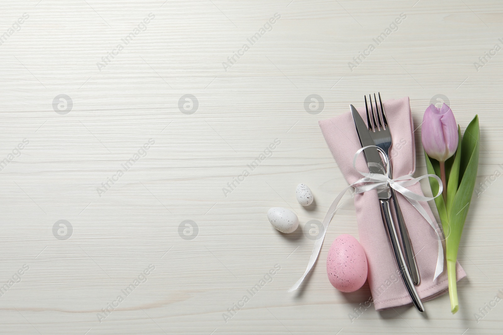 Photo of Cutlery set, painted eggs and beautiful flower on white wooden table, flat lay with space for text. Easter celebration