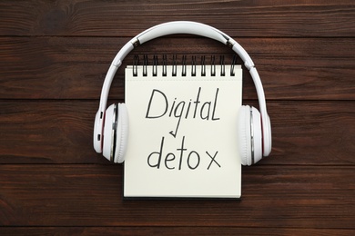 Photo of Notebook with phrase DIGITAL DETOX and headphones on wooden table, flat lay