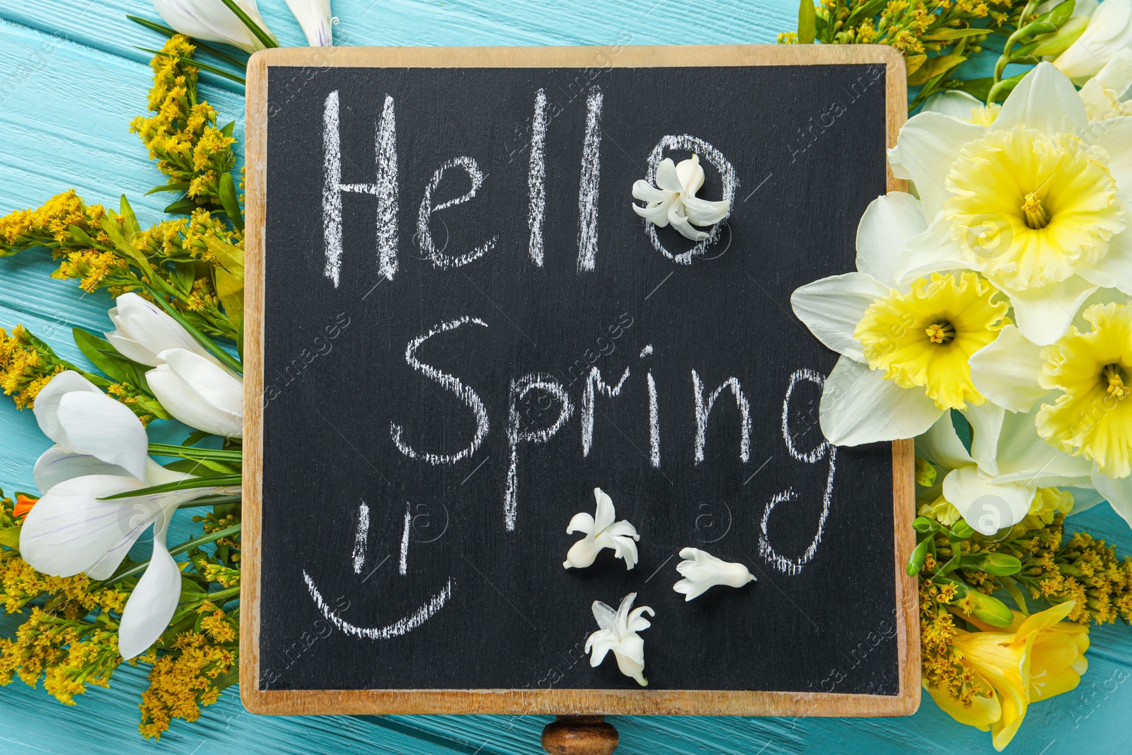 Photo of Blackboard with words HELLO SPRING and flowers on light blue wooden table, flat lay