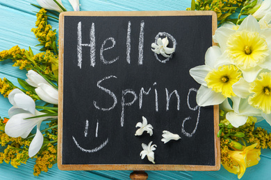 Blackboard with words HELLO SPRING and flowers on light blue wooden table, flat lay