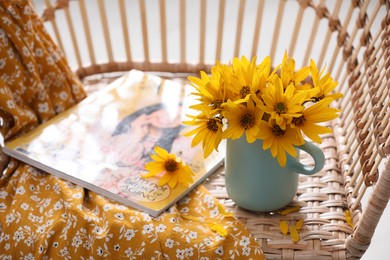 Beautiful bright flowers in light blue cup near magazine and fabric on rattan armchair. Space for text