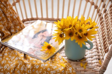 Photo of Beautiful bright flowers in light blue cup near magazine and fabric on rattan armchair. Space for text