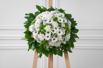 Photo of Funeral wreath of flowers on wooden stand near white wall indoors
