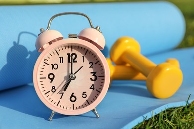 Photo of Alarm clock, dumbbells and fitness mat on green grass outdoors, closeup. Morning exercise