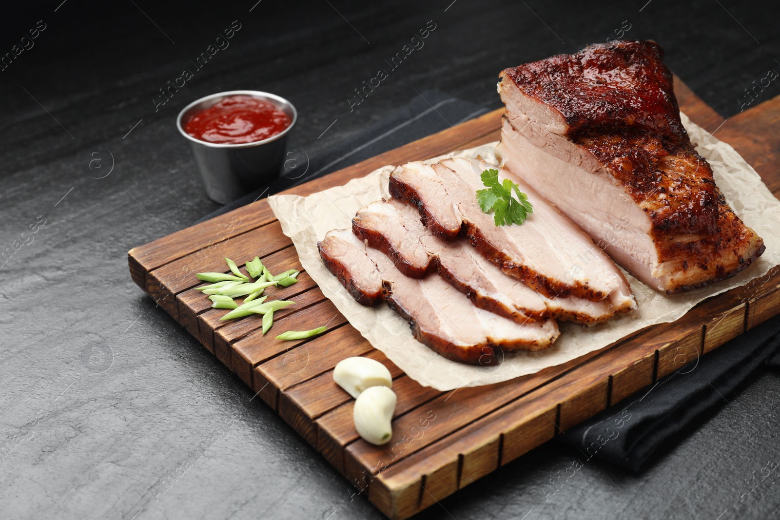 Photo of Pieces of baked pork belly served with sauce and parsley on black textured table