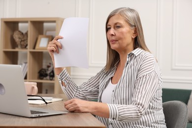 Photo of Menopause. Woman waving paper sheet to cool herself during hot flash at wooden table indoors