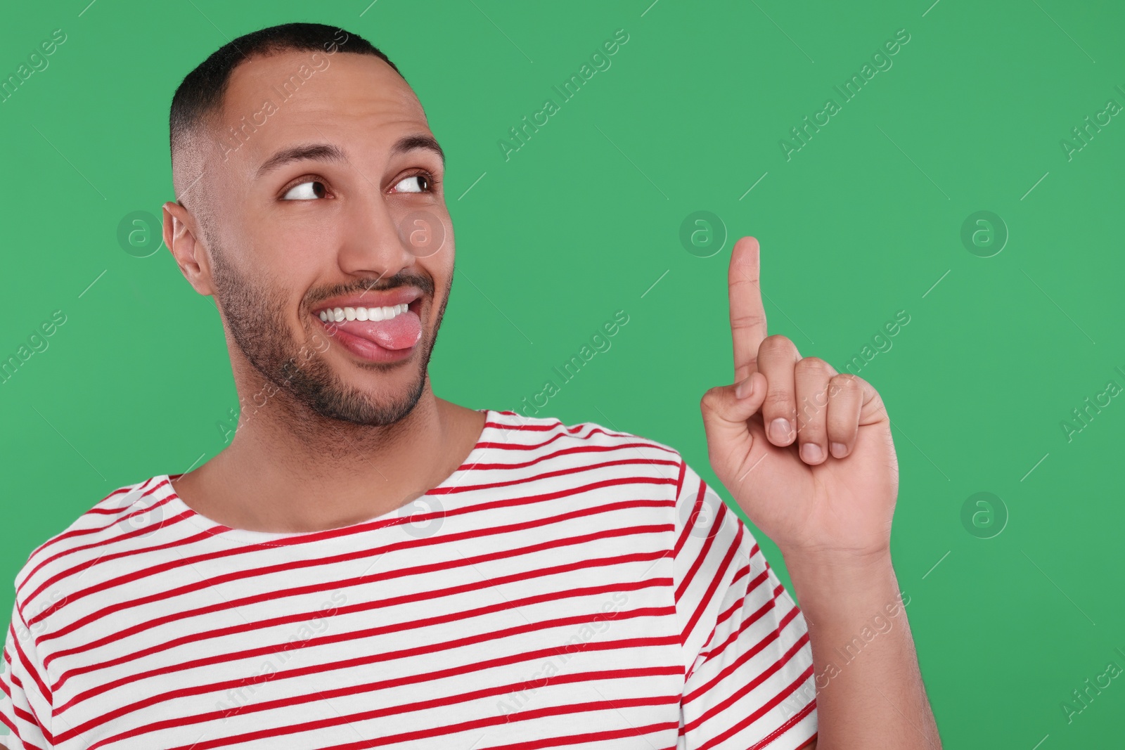 Photo of Happy young man showing his tongue and pointing at something on green background