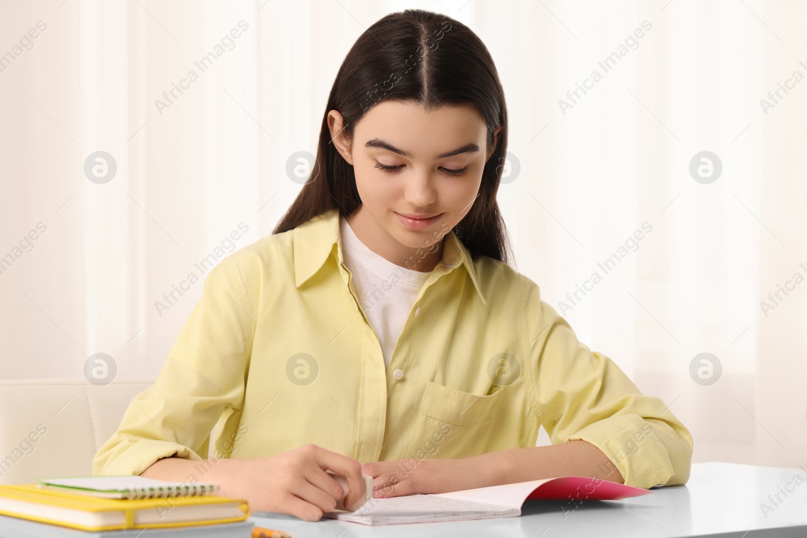 Photo of Teenage girl erasing mistake in her notebook at white desk indoors