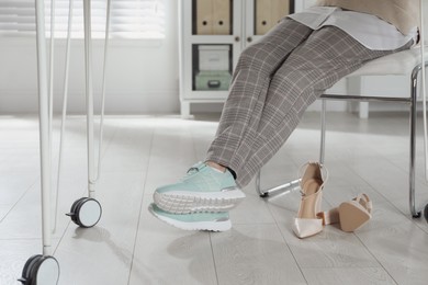 Photo of High heeled shoes near businesswoman wearing comfortable sneakers in office, closeup