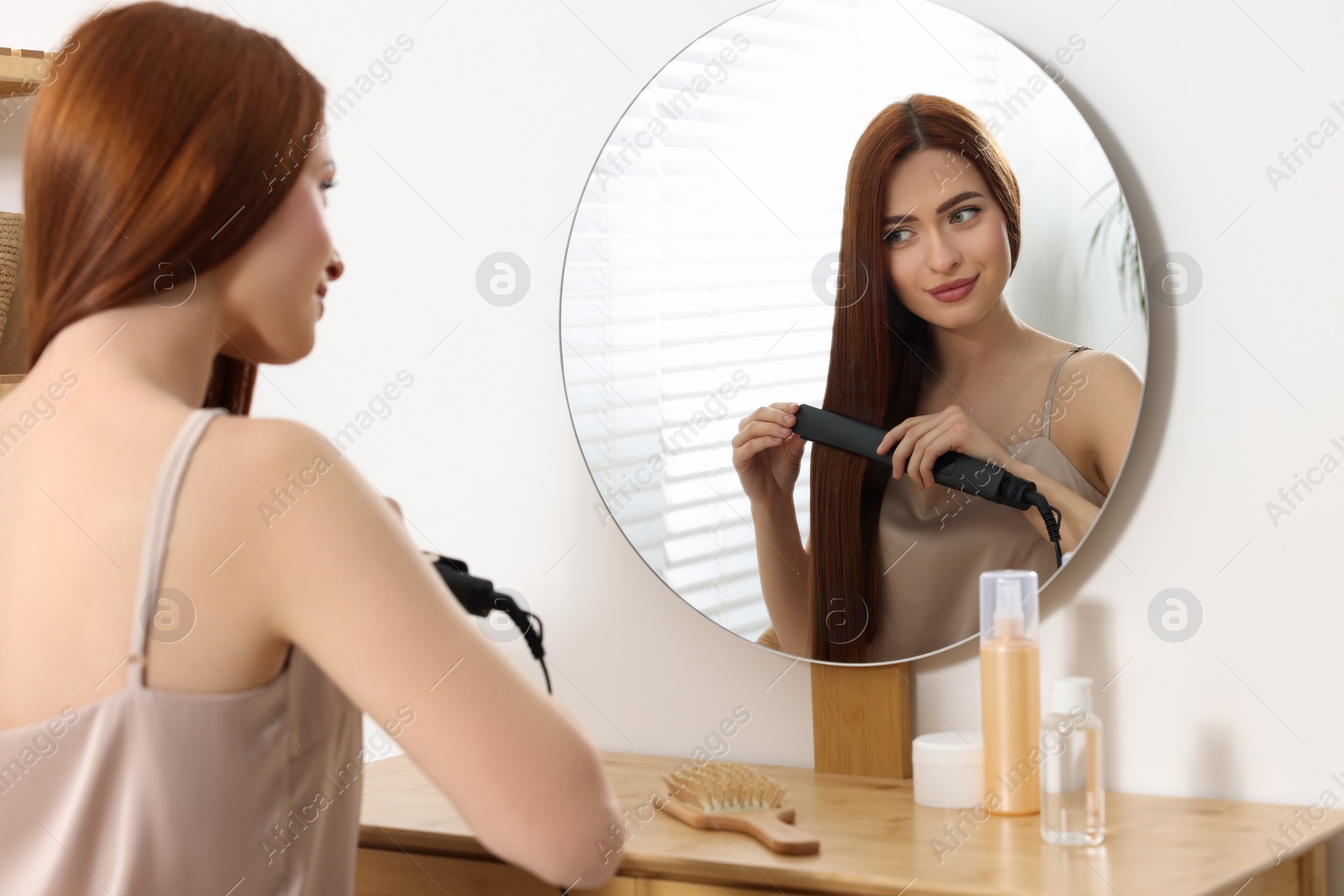 Photo of Beautiful woman using hair iron near mirror in room