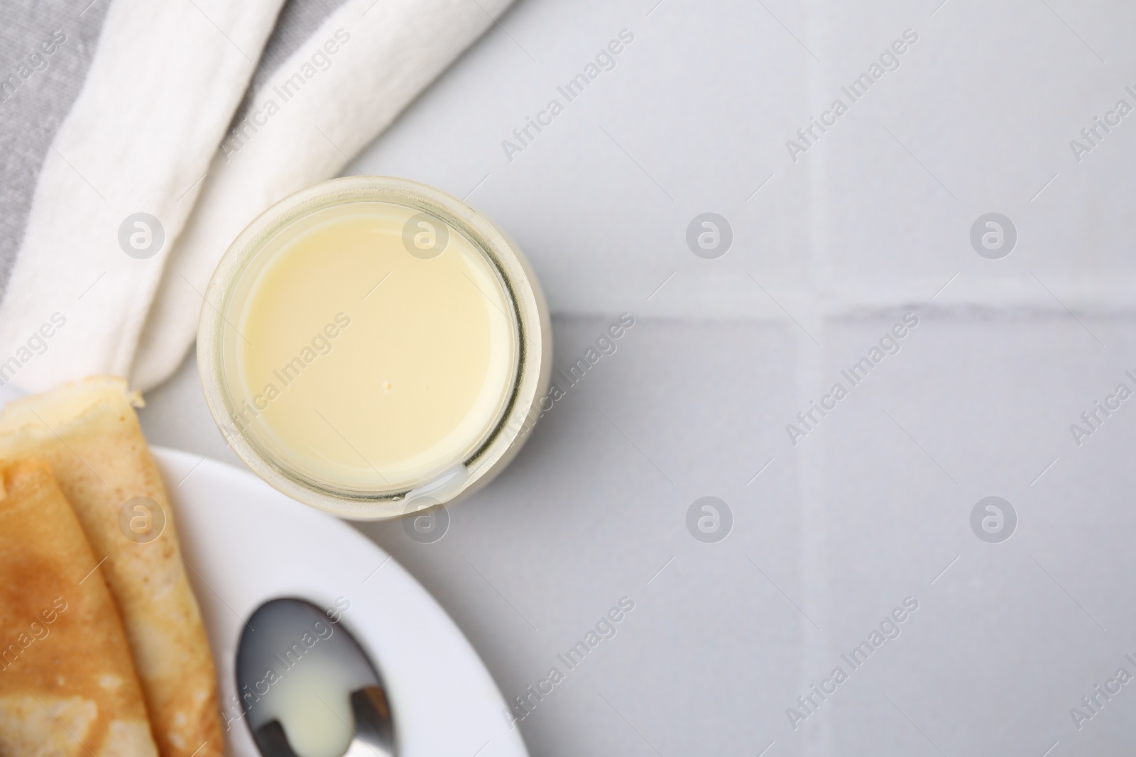 Photo of Tasty condensed milk in jar and crepes on white tiled table, flat lay. Space for text
