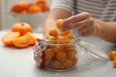 Woman putting yellow tomatoes into glass jar at light kitchen table, closeup. Pickling vegetables