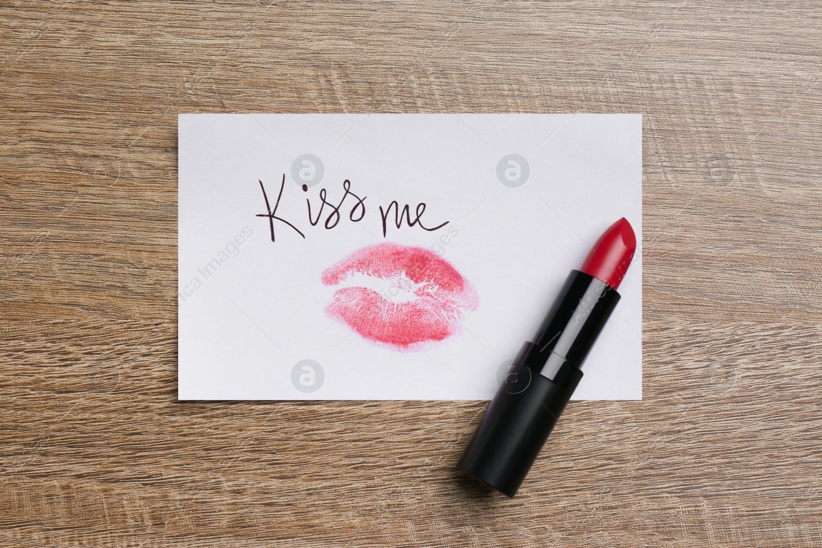Photo of Paper card with lip print, phrase Kiss Me and lipstick on wooden table, top view
